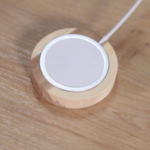 Peut inclure: Un chargeur sans fil rond avec une base en bois clair et un bord m&eacute;tallique argent&eacute;. La surface de chargement est blanche et un c&acirc;ble blanc s'&eacute;tend de l'appareil. Le chargeur est sur une surface en bois.