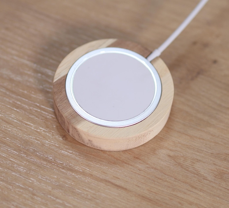 Peut inclure: Un chargeur sans fil rond avec une base en bois clair et un bord m&eacute;tallique argent&eacute;. La surface de chargement est blanche et un c&acirc;ble blanc s'&eacute;tend de l'appareil. Le chargeur est sur une surface en bois.