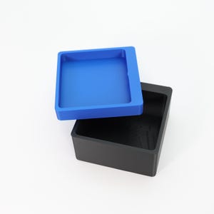 May include: A square, two-piece container with a blue lid and a black base. The lid is a lighter shade of blue and sits slightly above the black base. The container is empty and sits on a white surface.