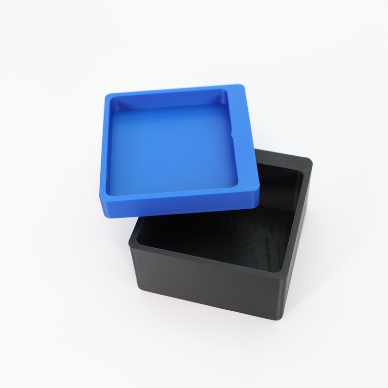 May include: A square, two-piece container with a blue lid and a black base. The lid is a lighter shade of blue and sits slightly above the black base. The container is empty and sits on a white surface.