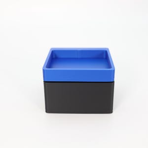 May include: A two-tone box with a square shape. The top is a vibrant blue, and the base is black. The box has a simple, modern design, suitable for storage or display. The box is made of a solid material.