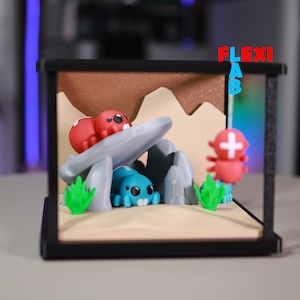 May include: A miniature diorama featuring a cave scene with cartoonish creatures. The scene includes red and blue figures, gray rocks, and green plants, all set against a tan and brown backdrop within a black frame. The word "FLEXI LAB" is visible.