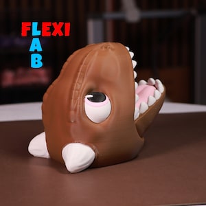 May include: A brown dinosaur-shaped toy with a large open mouth, white teeth, and a pink tongue. The toy has a white eye with a black pupil and a pink eyelid.