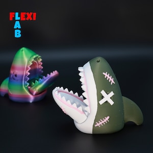 May include: Two 3D printed shark figurines with open mouths. One shark is green and white with pink stitching and a white cross on its head. The other shark is rainbow colored.