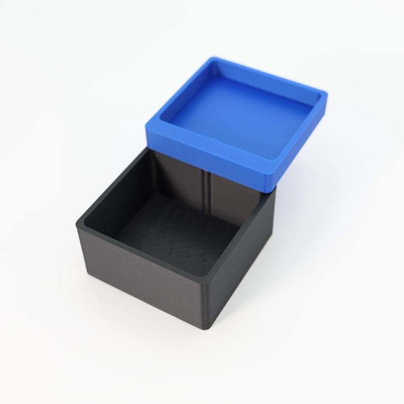 May include: A black square box with a blue lid. The lid is slightly ajar, revealing the interior of the box. The box and lid are made of a matte material. The background is white.
