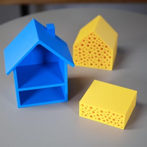 以下が含まれることがあります： 原色を使った3つのミニチュアハウスモデル。煙突とオープンシェルフが付いた青い家、穴あきデザインの黄色い家、穴の開いた黄色の長方形のブロック。オブジェクトは灰色の表面にあります。