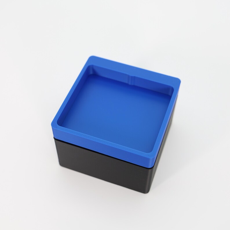 May include: A square, two-tone container with a black base and a bright blue top. The top has a raised rim, creating a tray-like design. The container is set against a plain white background.