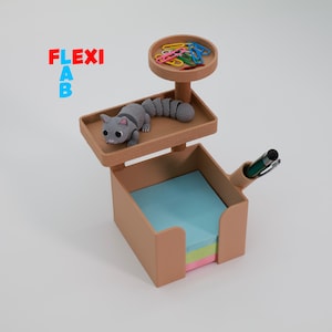 May include: A tan desk organizer with a cat figurine, a small tray with colorful paper clips, and a pen holder. The organizer holds a stack of sticky notes. The text "FLEXI AB" is in the upper left corner.