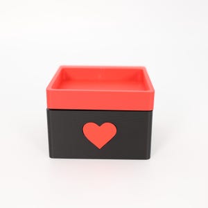K&ouml;nnte beinhalten: Kleine, quadratische Box mit roter Oberseite und schwarzer Basis. Eine rote Herzform ist aus der Vorderseite der Box ausgeschnitten. Die Box steht auf einer wei&szlig;en Oberfl&auml;che.