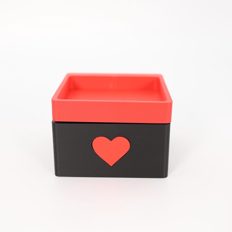 K&ouml;nnte beinhalten: Kleine, quadratische Box mit roter Oberseite und schwarzer Basis. Eine rote Herzform ist aus der Vorderseite der Box ausgeschnitten. Die Box steht auf einer wei&szlig;en Oberfl&auml;che.