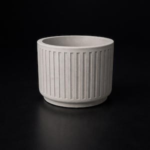 K&ouml;nnte beinhalten: Ein hellgrauer, zylindrischer Pflanztopf mit vertikalen Rippendetails. Der Pflanztopf hat einen abgerundeten Boden und einen glatten Innenraum. Die neutrale Farbe und das minimalistische Design eignen sich f&uuml;r verschiedene Pflanzen und Dekorationsstile.