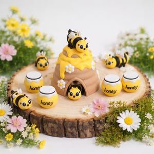 May include: A whimsical display featuring a beehive with a yellow honey drip, surrounded by small bee figurines and honey pots. The scene is set on a wooden slice, accented with flowers and greenery, creating a charming, nature-inspired theme.