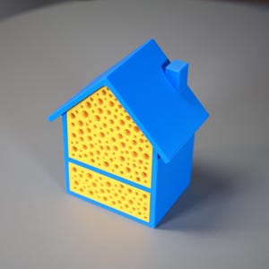 以下が含まれることがあります： 青と黄色の家型のオブジェクト。屋根と本体は青色で、壁は穴あき模様の黄色です。屋根には小さな煙突があります。このオブジェクトはおそらく装飾品または容器です。
