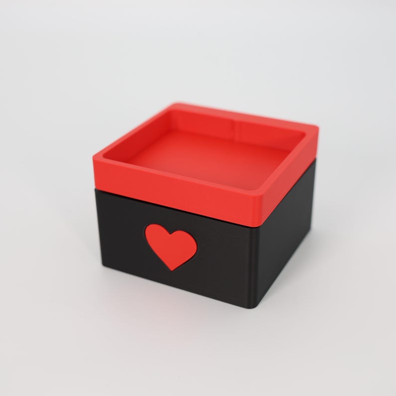 K&ouml;nnte beinhalten: Eine quadratische, zweifarbige Box mit rotem Deckel und schwarzer Basis. Ein rotes Herz ist aus der Vorderseite der schwarzen Basis ausgeschnitten. Die Box steht auf einer wei&szlig;en Oberfl&auml;che.