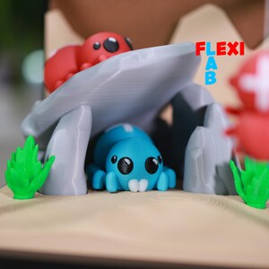 May include: A whimsical diorama featuring a blue spider-like creature under a gray stone structure, with a red creature on top. Green plant-like elements and a tan base complete the scene. The text "FLEXI AB" is visible.