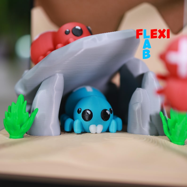 May include: A whimsical diorama featuring a blue spider-like creature under a gray stone structure, with a red creature on top. Green plant-like elements and a tan base complete the scene. The text "FLEXI AB" is visible.