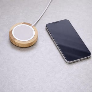 Peut inclure: Un chargeur sans fil en bois avec un centre blanc et un smartphone &agrave; cadre argent&eacute;. Le chargeur est rond et connect&eacute; &agrave; un c&acirc;ble blanc. Le t&eacute;l&eacute;phone est noir avec un cadre argent&eacute;. Les articles sont sur une surface grise.