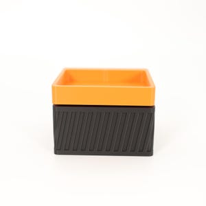 Puede incluir: Una caja de dos tonos con una parte superior cuadrada naranja y una base negra. La base negra tiene un dise&ntilde;o texturizado y acanalado. La caja est&aacute; sobre un fondo blanco.
