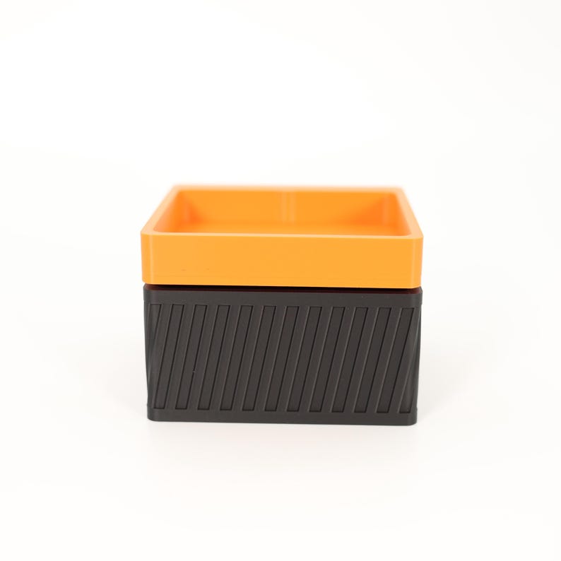 Puede incluir: Una caja de dos tonos con una parte superior cuadrada naranja y una base negra. La base negra tiene un dise&ntilde;o texturizado y acanalado. La caja est&aacute; sobre un fondo blanco.