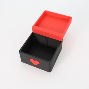 K&ouml;nnte beinhalten: Eine schwarze Box mit rotem Deckel, leicht ge&ouml;ffnet. Die Box hat ein rotes Herz auf der Vorderseite. Der quadratische Deckel passt gut oben drauf. Die leere Box steht auf einer wei&szlig;en Oberfl&auml;che.