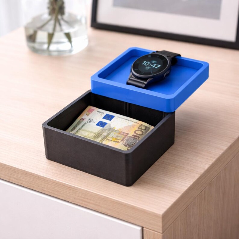 May include: A black and blue box with a watch and money inside. The watch has a black band and a digital display showing the time. The box contains a 100 Euro bill. The box is sitting on a wooden surface.
