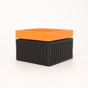 Puede incluir: Una caja de almacenamiento geom&eacute;trica de dos piezas. La parte superior es un cuadrado naranja brillante, y la base es un cuadrado negro con l&iacute;neas verticales en &aacute;ngulo. La caja est&aacute; sobre un fondo blanco.
