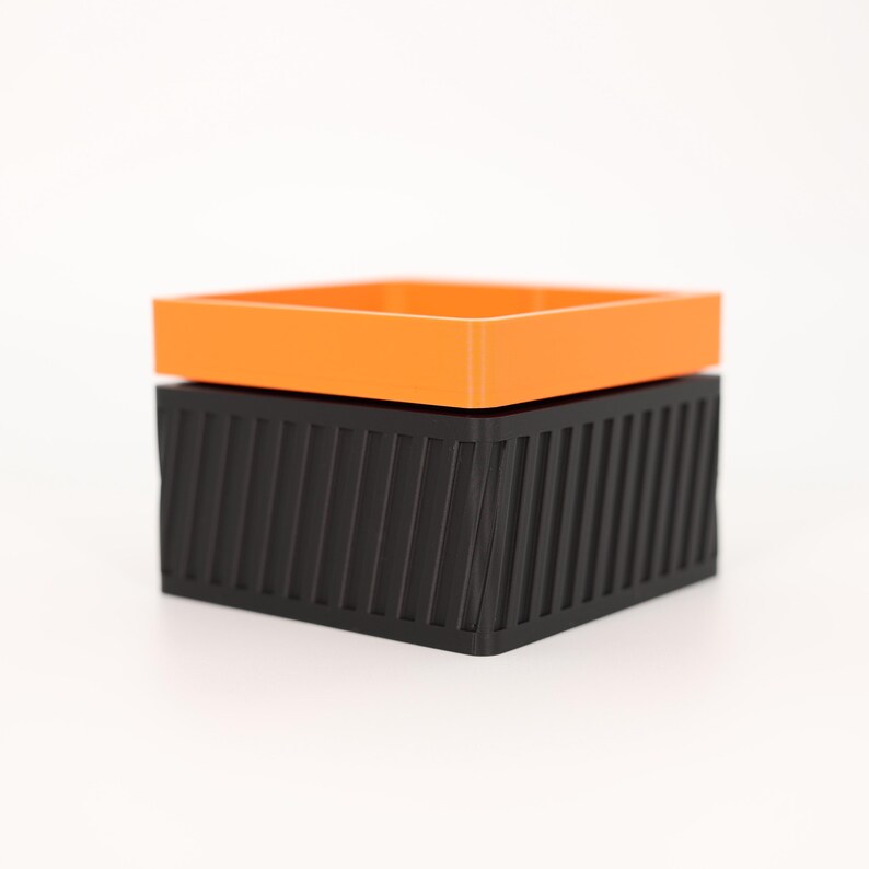 Puede incluir: Una caja de almacenamiento geom&eacute;trica de dos piezas. La parte superior es un cuadrado naranja brillante, y la base es un cuadrado negro con l&iacute;neas verticales en &aacute;ngulo. La caja est&aacute; sobre un fondo blanco.