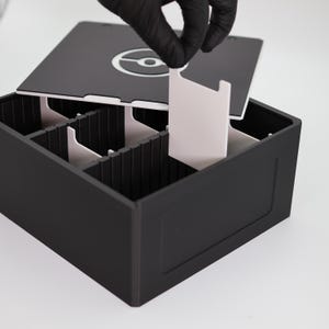 Könnte beinhalten: Eine schwarze Aufbewahrungsbox mit weißem Innenraum und Trennwänden. Der Deckel hat eine weiße Grafik. Eine Hand in einem schwarzen Handschuh hebt eine weiße Trennwand aus der Box. Die Box ist zum Organisieren von Gegenständen konzipiert.