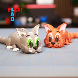 May include: Two cartoon cat figurines, one gray with green eyes and one orange with brown eyes. Both cats are lying down with their heads resting on the surface. The cats are made of plastic and have a cartoonish style.