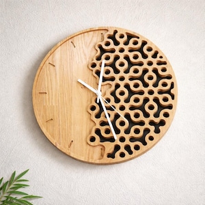 K&ouml;nnte beinhalten: Eine runde Wanduhr aus Holz mit einem einzigartigen Design. Das Zifferblatt ist geteilt, mit einem Massivholzteil auf der linken Seite und einem gemusterten, durchbrochenen Design auf der rechten Seite. Wei&szlig;e Zeiger zeigen die Uhrzeit an. Die Uhr ist an einer wei&szlig;en Wand befestigt.