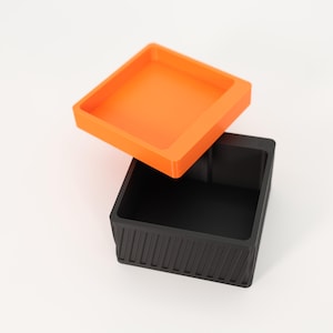 Puede incluir: Un recipiente cuadrado de dos piezas. La tapa es de color naranja brillante y la base es negra con detalles de estr&iacute;as verticales. La tapa est&aacute; ligeramente abierta, revelando el interior del recipiente.