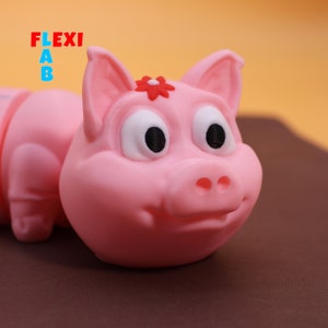 Peut inclure: Un jouet en caoutchouc rose en forme de cochon avec une fleur rouge sur la tête et de grands yeux noirs. Le cochon est couché sur le ventre, les pattes tendues.