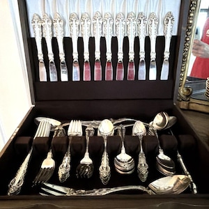 May include: A silver flatware set arranged in a dark wooden storage chest. The set includes twelve knives with ornate handles stored in the lid, and various forks and spoons organized in the velvet-lined base with dedicated slots.