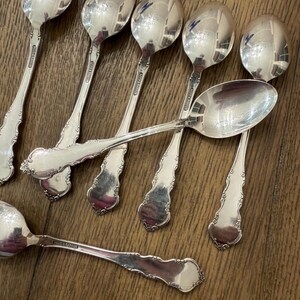 May include: A collection of silver spoons with ornate handles and reflective bowls. The spoons are arranged on a wooden surface, showcasing their polished finish. The spoons have the text "Reed & Barton" on the handle.