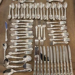 May include: A large collection of silver-toned flatware including numerous spoons, forks, and knives. The set features ornate, textured handles with intricate detailing. The pieces are arranged in organized rows and columns on a wooden surface.