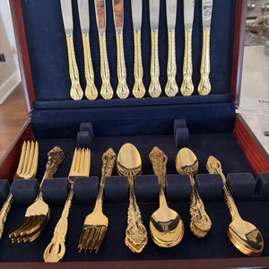 May include: A collection of gold-coloured cutlery, including knives, forks, and spoons, presented in a wooden case with a blue velvet lining. The silverware features ornate, detailed handles.