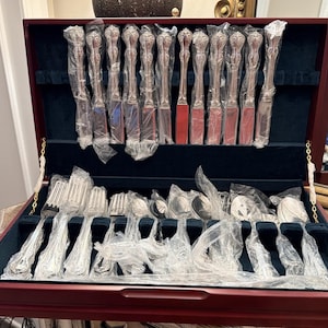 May include: A set of silver-toned flatware arranged in a dark wood storage chest with a navy blue velvet interior. The collection includes knives, forks, and spoons, each individually wrapped in clear plastic protective sleeves.