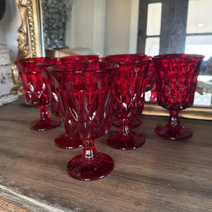 May include: A set of six ruby red vintage wine glasses. Each glass features a diamond-cut pattern on the bowl and a stemmed base. The glasses are arranged on a wooden surface, reflecting light and adding a touch of elegance.