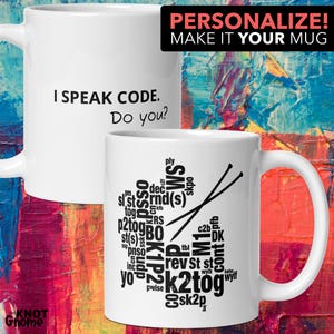 Op de afbeelding: Witte keramische mok met een zwart woordwolkontwerp met breistermen en een paar breinaalden. De tekst op de mok luidt "Ik spreek code. En jij?"