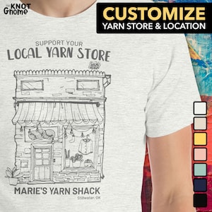 May include: A grey t-shirt featuring a black and white illustration of a yarn shop with the text "Support Your Local Yarn Store" and "Marie's Yarn Shack Stillwater, OK". The t-shirt can be customised with your own yarn shop name and location.