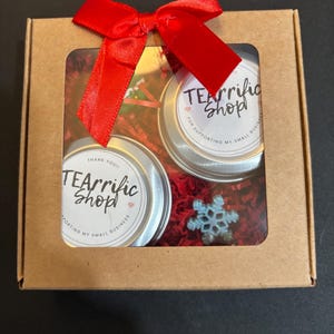 May include: A gift box with a red bow, containing two small tins with the text "TEArrific Shop" and a snowflake. The box is brown with a clear window, filled with red shredded paper.
