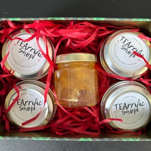 May include: A gift box containing four small, round tins and a small jar of honey. The tins have lids with the text "TEArrific Shop" and "Thank you for supporting my small business." Red paper shreds fill the box.