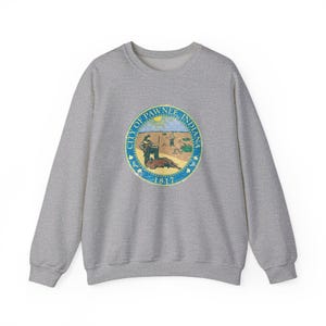 May include: A gray sweatshirt featuring a circular emblem with the text "City of Pawnee Indiana" and the year "1817". The emblem depicts a scene with figures and a beaver. The sweatshirt has a crew neck and long sleeves.