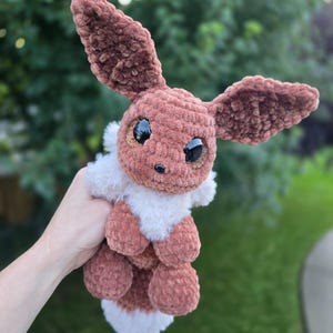 May include: A brown and white crocheted plush toy featuring large pointed ears, a fluffy white chest ruff, and oversized black eyes with glittery copper-colored irises. The creature has a small black nose and a soft, textured yarn construction.