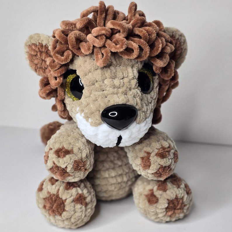 Pode incluir: Um brinquedo de pel&uacute;cia de le&atilde;o de croch&ecirc; feito &agrave; m&atilde;o. O le&atilde;o tem um corpo bege, uma juba marrom e manchas marrons nas patas. Possui olhos grandes e pretos com detalhes dourados brilhantes e um focinho branco. O brinquedo de pel&uacute;cia &eacute; macio e fofinho.
