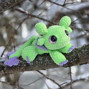 Puede incluir: Una criatura hecha a mano, verde, tejida a ganchillo, con grandes ojos brillantes y detalles morados en los pies y el cuello. El peluche est&aacute; posado en una rama contra un fondo nevado y borroso. La criatura tiene un dise&ntilde;o caprichoso y de dibujos animados.