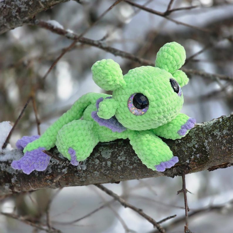 Puede incluir: Una criatura hecha a mano, verde, tejida a ganchillo, con grandes ojos brillantes y detalles morados en los pies y el cuello. El peluche est&aacute; posado en una rama contra un fondo nevado y borroso. La criatura tiene un dise&ntilde;o caprichoso y de dibujos animados.