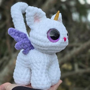 May include: White crocheted cat with a purple wing, a gold horn, and large pink and purple eyes.