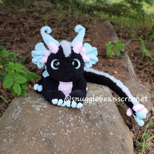 May include: A black and pink crocheted dragon plush toy with blue accents. The dragon is sitting on a rock with its tail curled around the rock. The dragon has large eyes and a friendly expression.