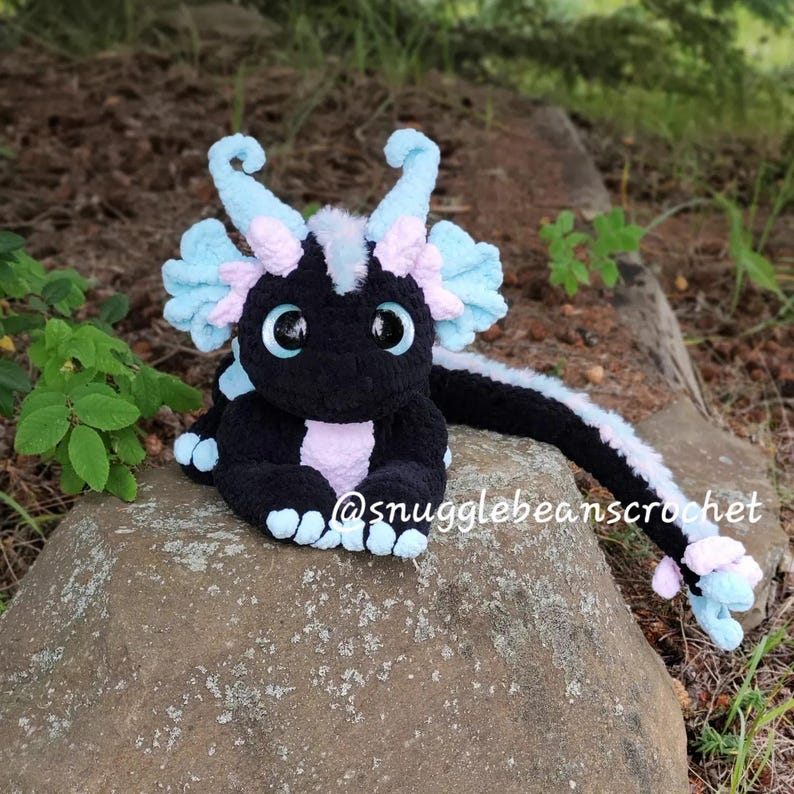 May include: A black and pink crocheted dragon plush toy with blue accents. The dragon is sitting on a rock with its tail curled around the rock. The dragon has large eyes and a friendly expression.